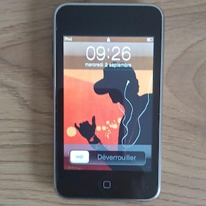 May include: A silver and black digital media player with a rectangular touchscreen display. The screen shows the time 09:26, the date mercredi 2 septembre, and a graphic of a silhouette wearing headphones. The unlock slider displays the text Déverrouiller.