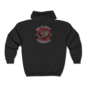 May include: A black zip-up hoodie featuring a graphic on the back. The design shows a grey security camera inside a red circle with a diagonal line through it. The text DE-FLOCK AMERICA is printed in grey around the circle with two small stars.