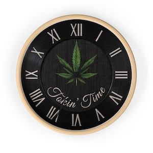 Op de afbeelding: Een ronde wandklok met een lichthouten frame en een zwarte wijzerplaat met een groene cannabisblad-afbeelding in het midden. De klok heeft witte Romeinse cijfers en de tekst "Tokin' Time" in een wit cursief lettertype onder het blad.