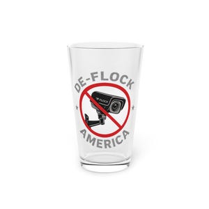 May include: A clear glass pint glass featuring a graphic of a black security camera crossed out by a red circle. The text DE-FLOCK AMERICA is printed in grey capital letters around the graphic, with two small grey stars on the sides.