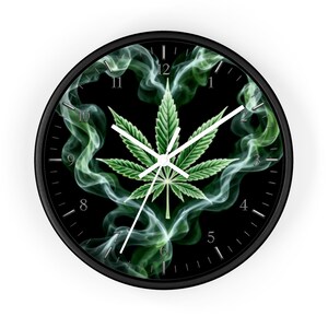 Pode incluir: Um relógio de parede redondo com moldura preta e mostrador preto apresentando um gráfico de folha de cannabis verde cercado por padrões de fumaça branca e verde. O relógio possui ponteiros brancos de horas, minutos e segundos e números brancos.