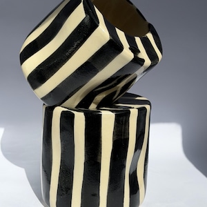 May include: Two cylindrical ceramic cups stacked vertically, featuring a hand-painted pattern of bold, vertical black stripes against a cream-colored background. The cups have a glossy finish and slightly irregular, organic shapes.