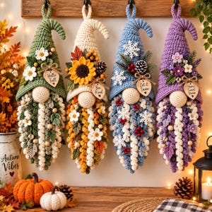 May include: Four hanging crochet gnome decorations with pointed hats, each featuring unique seasonal embellishments like flowers, pinecones, and snowflakes. Each gnome has a wooden heart tag with text reading Hello Fall, Bee Kind, Let it Snow, and Home Sweet Home.