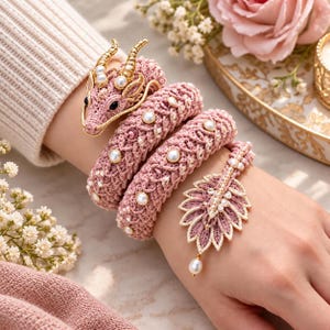 May include: A wrap-around bracelet featuring a crocheted pink dragon head with gold-toned horns and black eyes, a long coiled body embellished with white pearls, and a leaf-shaped terminal adorned with pearls and a single dangling pearl drop.