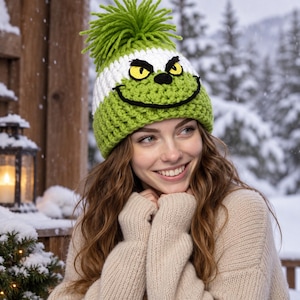 May include: A lime green and white crocheted beanie featuring a large lime green yarn pom-pom on top. The hat displays a character face with yellow eyes, black pupils, and a wide black stitched smile across the front of the green section.