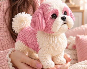 May include: A small, handcrafted crochet dog figurine featuring a pink and cream colour scheme. The plush toy has a textured, yarn-based coat, large dark eyes, a black nose, and a fluffy, upright tail. The design mimics a Shih Tzu breed.