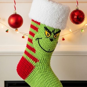 Peut inclure: Une chaussette de Noël crochetée avec une base verte, des rayures horizontales rouges sur le côté et un revers blanc duveteux. L'avant présente un visage brodé avec des yeux jaunes, des sourcils noirs et un large sourire noir courbé.
