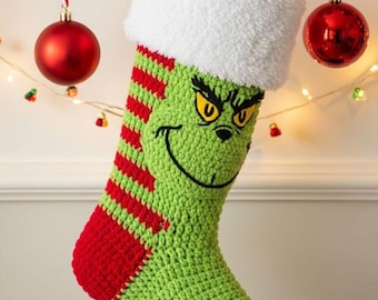May include: A crocheted Christmas stocking featuring a green base with red horizontal stripes on the side and a white fuzzy cuff. The front displays an embroidered face with yellow eyes, black eyebrows, and a wide, curved black smile.