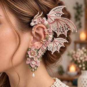 May include: A crochet ear cuff featuring a dragon design with cream and pink wings, accented by small white beads. The lower section consists of layered crochet flowers in shades of pink and green, finished with a single dangling white pearl bead.