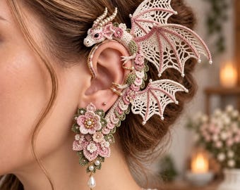 May include: A crochet ear cuff featuring a dragon design with cream and pink wings, accented by small white beads. The lower section consists of layered crochet flowers in shades of pink and green, finished with a single dangling white pearl bead.