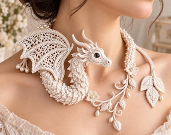 May include: A cream-coloured crochet dragon necklace featuring intricate lace wings, a textured body, and small pearl accents. The design includes delicate floral motifs, vine-like extensions, and leaf-shaped ends, creating a detailed statement piece.