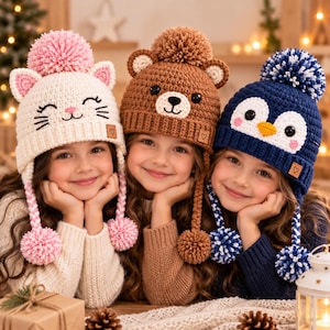 Op de afbeelding: Drie gehaakte mutsen met dierendesigns: een witte kat met roze oren en pompons, een bruine beer met ronde oren en een pompon, en een donkerblauwe pinguïn met een wit gezicht, gele snavel en een blauw-witte pompon.