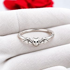 May include: A silver-toned metal ring featuring a small bat motif at the center. The bat has outstretched wings and two tiny black circular accents representing eyes. The band is thin, smooth, and circular, designed to sit flat against a finger.