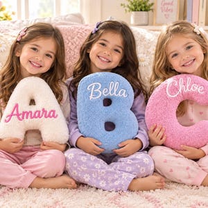 May include: Three plush letter-shaped pillows in white, blue, and pink. The white pillow is shaped like the letter A with the name Amara embroidered in pink. The blue pillow is shaped like the letter B with the name Bella in white. The pink pillow is shaped like the letter C with the name Chloe in white.