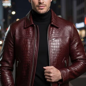 Peut inclure: Une veste bordeaux profond avec un motif texturé en peau de crocodile, un col classique et des fermetures éclair en métal argenté sur le devant et les poches latérales. Le vêtement est porté sur un pull noir à col montant et manches longues.
