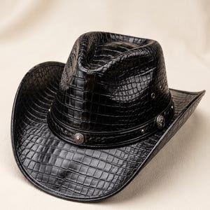 Op de afbeelding: Een zwarte cowboyhoed met een glanzende krokodillenleertextuur over de gehele kroon en rand. De hoed is voorzien van een decoratieve zwarte band versierd met ronde zilverkleurige metalen concho's en kleine metalen oogjes aan de zijkanten.