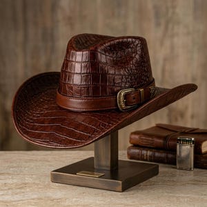 May include: A dark brown cowboy hat with a textured crocodile skin pattern. The hat features a smooth brown leather band around the crown, secured by an ornate, antique-style brass buckle. The hat is displayed on a wooden stand.