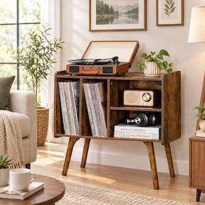 May include: A dark wood record player stand with four angled legs, featuring two vertical storage compartments filled with vinyl records, a shelf holding a vintage-style radio, black headphones, and two books labeled Radio & Turntable and Cam Westmeetseast.