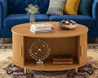 May include: A round light wood coffee table with vertical slatted sliding doors, gold handles, and short gold legs. The table features an open center shelf containing a metal solar system model and two books, with three books and a bowl of chocolates on top.