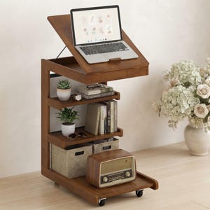 May include: A brown wooden rolling side table with an adjustable tilting top surface holding a silver laptop. The unit features three open shelves containing two potted green plants, several books, two fabric storage bins, and a vintage radio.