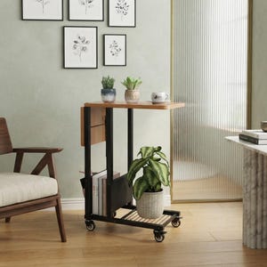 May include: A mobile side table with a light wood top and a black metal frame on four casters. The unit features a lower slatted shelf, a vertical storage compartment holding several books, and a top surface holding a small potted plant and a cup.
