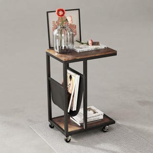 May include: A C-shaped side table with a dark wood-grain top and bottom shelf, supported by a black metal frame on four casters. It features a black fabric magazine holder, a glass vase with artificial flowers, and several books on the surfaces.