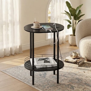 May include: A round black side table with three thin legs and a lower shelf featuring a metal railing. The tabletop includes a built-in power strip with a charging cable, a white ceramic mug on a saucer, and a smartphone resting on the surface.