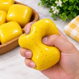 May include: A bright yellow, squishy, rounded stress relief toy with small red speckles throughout its surface. The object is being squeezed, showing its flexible, malleable texture. Other similar yellow items are visible in a wooden bowl nearby.