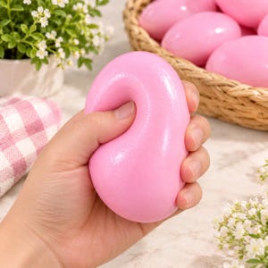 May include: A smooth, oval-shaped pink stress relief ball with a glossy finish. The object is pliable and shows a slight indentation from pressure, demonstrating its soft, squeezable texture. It is a solid, uniform light pink color throughout.