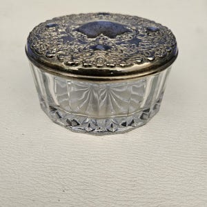 May include: A round glass trinket box with a clear, faceted base and a decorative silver-toned metal lid. The lid features an intricate, embossed floral pattern with a dark, recessed center, while the glass base has vertical cut grooves.