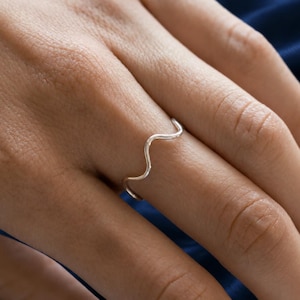 May include: A thin, polished silver ring featuring a single, gentle wave-like curve along its band. The metal has a smooth, reflective surface that catches the light, creating a simple and continuous undulating shape that encircles the finger.