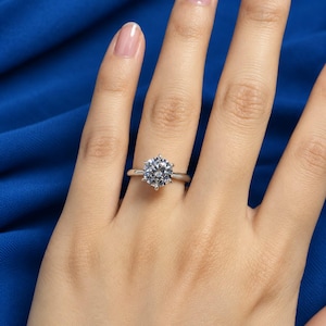 May include: A solitaire ring featuring a round brilliant-cut clear gemstone set in a polished silver-toned metal band with a six-prong setting. The ring is centered on the finger, showcasing the reflective facets of the stone and the smooth band.