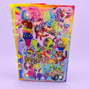 May include: A clear acrylic binder featuring a vibrant Lisa Frank graphic print. The cover displays colorful illustrations of animals, aliens, and rainbows, including a teddy bear, a tiger cub, and a car. The binder has white plastic rings on the side.