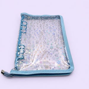 May include: A clear, six-ring binder cover featuring a holographic star pattern throughout the transparent material. The binder is edged with light blue fabric trim and includes a matching light blue zipper closure with a silver metal pull tab.