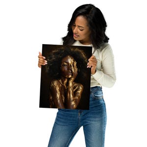 May include: A square photographic print featuring a portrait of a person with dark skin and voluminous dark hair. The subject is covered in metallic gold paint across their face, neck, and shoulders, with one hand partially covering their face.