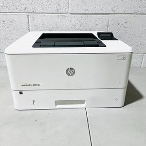 Pode incluir: Uma impressora a laser HP LaserJet Pro 4001dn branca com uma bandeja de saída superior preta, um pequeno visor digital e um botão de energia no painel frontal. O dispositivo inclui duas bandejas de papel rotuladas com os números 1 e 2.