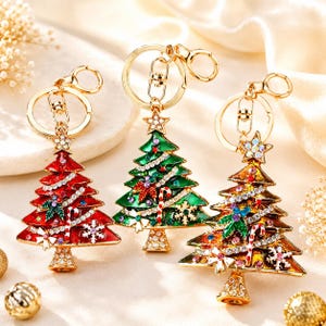 May include: Three metal Christmas tree keychains with gold-tone hardware. Each tree features enamel coloring in red, green, or multicolored metallic finishes, accented with clear rhinestones, small candy cane motifs, and snowflake details.