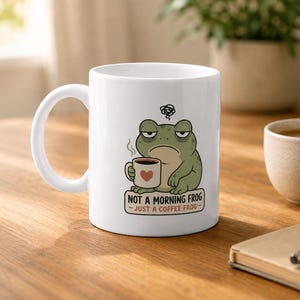 May include: A white ceramic mug featuring a cartoon illustration of a tired green frog holding a small mug with a heart design. Above the frog is a scribble symbol, and below it is the text: NOT A MORNING FROG - JUST A COFFEE FROG.