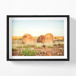 May include: A framed landscape photograph featuring large, rounded orange-brown boulders situated in a dry, grassy field under a pale blue sky. The scene captures a desert-like terrain with sparse vegetation and distant hills in the background.