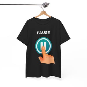 May include: A black short-sleeved t-shirt featuring the word PAUSE in white capital letters above a teal circular icon with two vertical white bars. A graphic of a hand is shown pressing the pause button on the front of the garment.