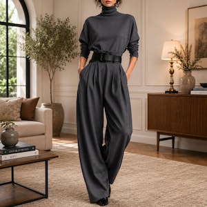 May include: A dark charcoal gray jumpsuit featuring a high turtleneck, long sleeves, and wide-leg trousers with deep pleats at the waist. The garment is cinched with a wide black leather belt that has a prominent rectangular buckle.