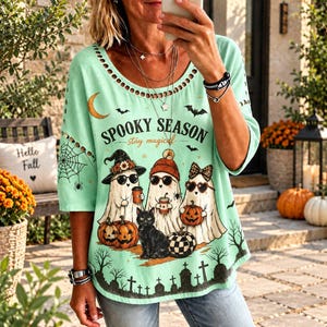 May include: A light green t-shirt with a round neckline featuring cut-out details. The graphic print displays three ghosts wearing sunglasses and hats, a black cat, pumpkins, bats, a crescent moon, and the text SPOOKY SEASON stay magical.