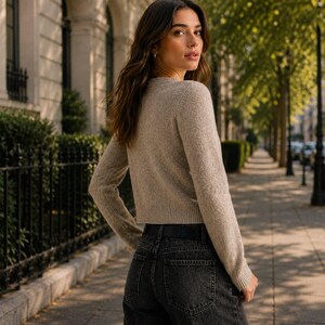 May include: A long-sleeved, beige, crew-neck sweater with a textured knit finish, paired with black denim jeans featuring a black leather belt. The sweater has a cropped length with a ribbed hem and cuffs, while the jeans have a classic five-pocket design.