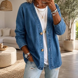 Könnte beinhalten: Eine mittelblaue Jeans-Strickjacke mit V-Ausschnitt, langen Ärmeln und zwei großen braunen Knöpfen auf der Vorderseite. Das Kleidungsstück wird über einem schlichten weißen T-Shirt getragen und mit einer hellblauen Jeans mit Used-Details kombiniert.