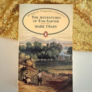 Op de afbeelding: Een paperbackboek getiteld The Adventures of Tom Sawyer van Mark Twain, onderdeel van de Penguin Popular Classics-serie. De omslag bevat een landschapsillustratie van een landelijk tafereel met twee figuren, glooiende heuvels en een rivier.