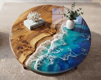 May include: A round coffee table featuring a natural wood slab on one side and a glossy blue resin pour on the other, mimicking ocean waves with white foam details. A stack of books, a white coral sculpture, and a vase with greenery sit on top.