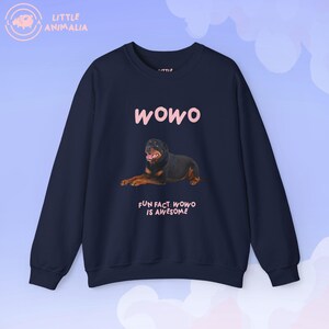 Puede incluir: Una sudadera de manga larga en color azul marino oscuro con un gráfico de un perro Rottweiler tumbado. Sobre el perro aparece el texto "WOWO" en rosa claro, y debajo del perro el texto "FUN FACT: WOWO IS AWESOME" en letras rosa claro.
