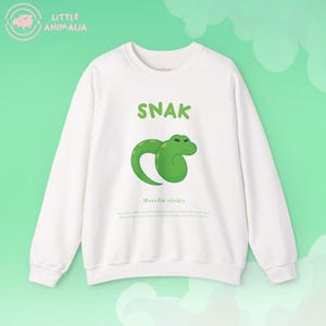 May include: A white crewneck sweatshirt featuring a green cartoon snake illustration in the center. Above the snake, the text reads "SNAK" in green letters. Below the snake, the scientific name "Morelia viridis" is printed in small text.