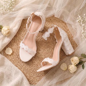 May include: A pair of white block-heeled sandals featuring an ankle strap and a toe strap adorned with white floral lace and small pearls. The heel is covered in a textured white fabric with pearl accents, and the insole is a light beige color.