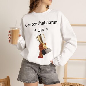 May include: A white long-sleeve sweatshirt featuring the black text Center that damn < div > above a graphic of a hand holding a gold trophy. The sweatshirt is paired with gray denim shorts, and a clear plastic cup with a straw is held nearby.